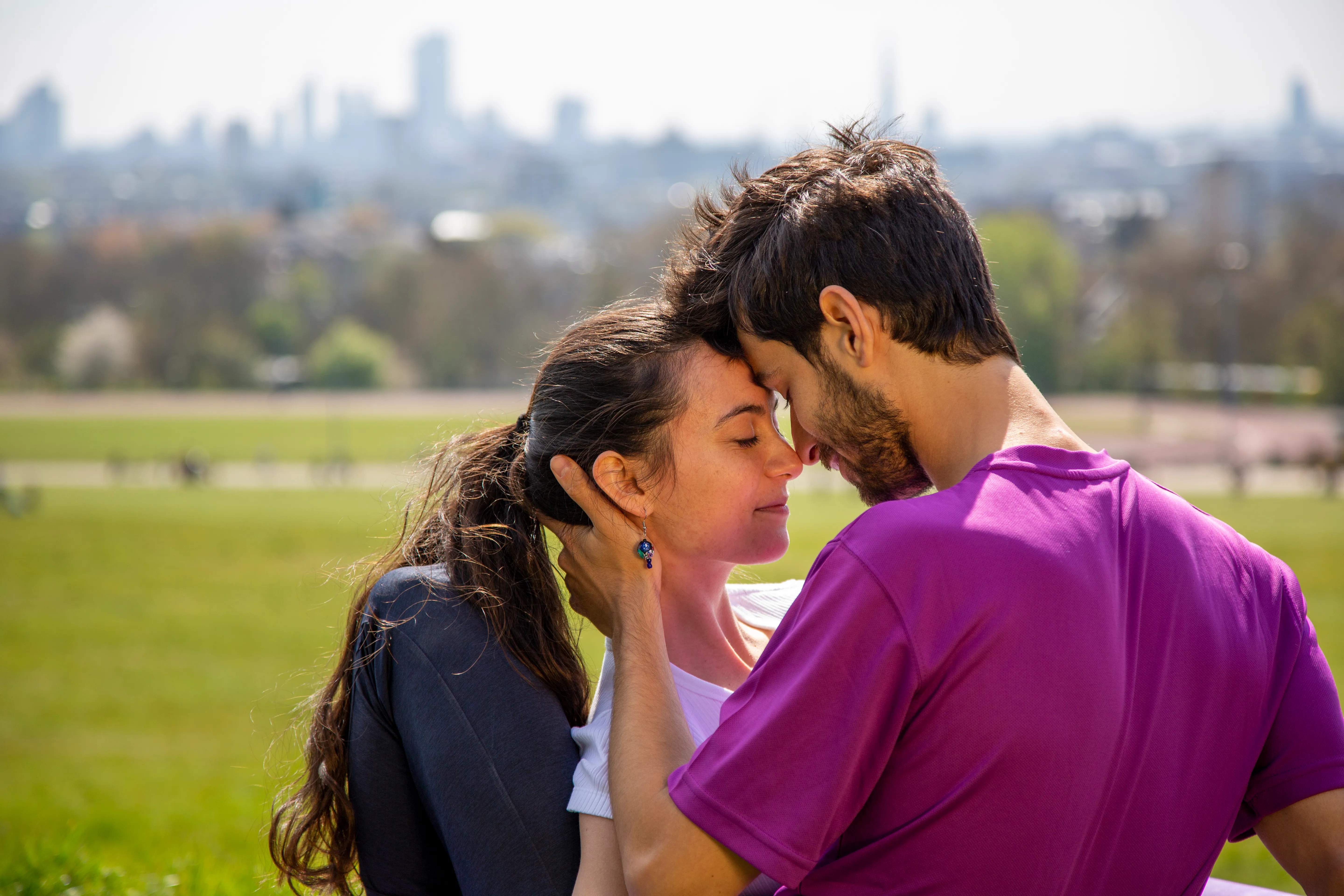 A couple sitting together in a park with the London skyline behind them